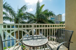 A balcony with a round table and two chairs, overlooking a body of water and palm trees.