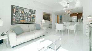 Uma sala de estar moderna com um sofá branco, uma mesa de centro de vidro e uma grande pintura com vista para a paisagem urbana.