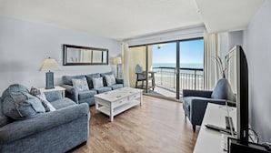 En moderne stue med sofa, sofabord og fjernsyn, en balkon med havudsigt og et spiseområde med stole.