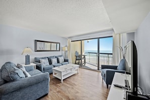 Relax in this stylish living room with a large flat-screen TV, comfortable sofa, and sliding glass door leading to a balcony with ocean views.