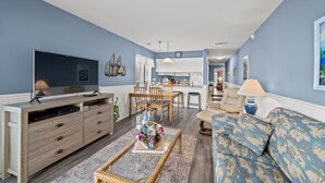 Un salon avec un téléviseur, un canapé et une table basse. On y trouve également un coin repas avec des chaises, et une cuisine à l  ’arrière-plan.