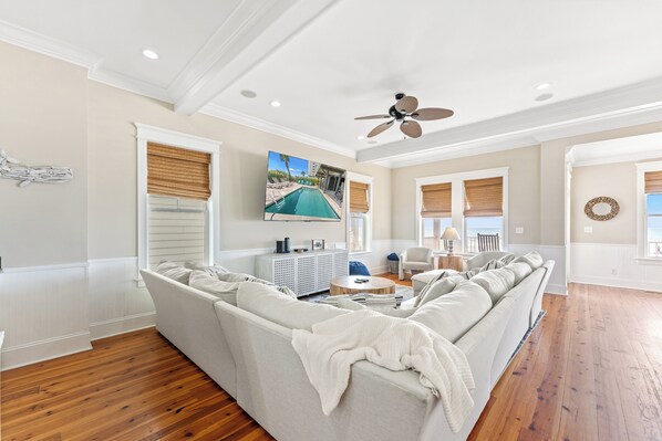 A living room with a sectional sofa, a ceiling fan, a TV mounted on the wall, and a view of a pool through the window.
