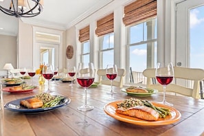 Mesa de jantar posta com pratos de comida e copos de vinho, janelas amplas com persianas e um lustre.