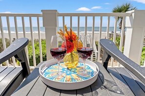 A table with a decorative tray, two wine glasses, and a vase with flowers on a balcony overlooking the beach.