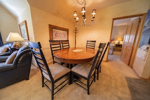 A dining area with a round wooden table, six chairs, a chandelier, and a sofa with cushions.