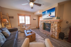A living room with a stone fireplace, a television mounted above it, a dark leather sofa, a wooden coffee table, and a ceiling fan. - Expedition Station 8608 (Keystone)