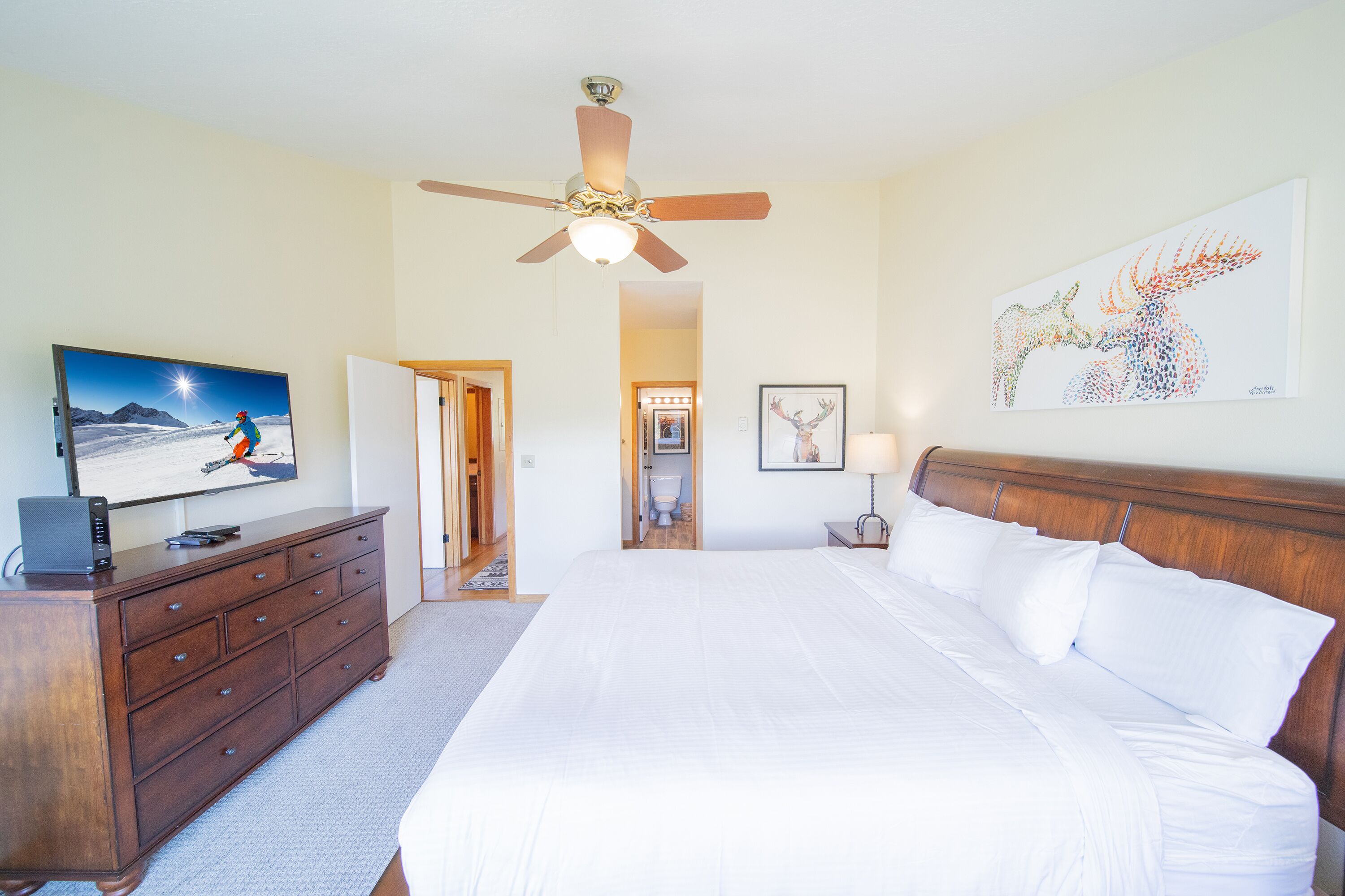A bedroom with a large bed, a ceiling fan, a wooden dresser, a television, and artwork on the walls.