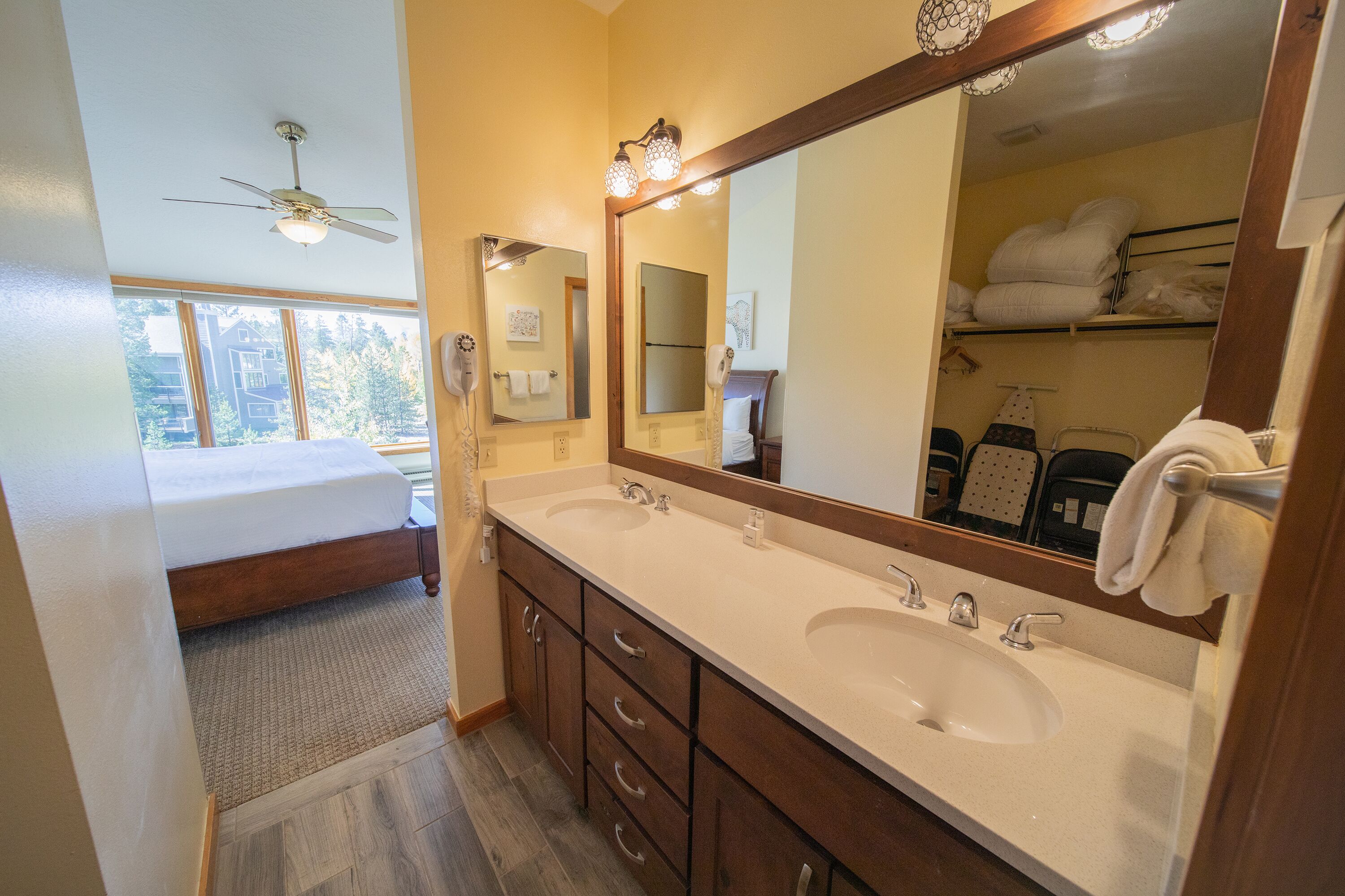 A bathroom with a double sink, a large mirror, a ceiling fan, and a bed with a window view.