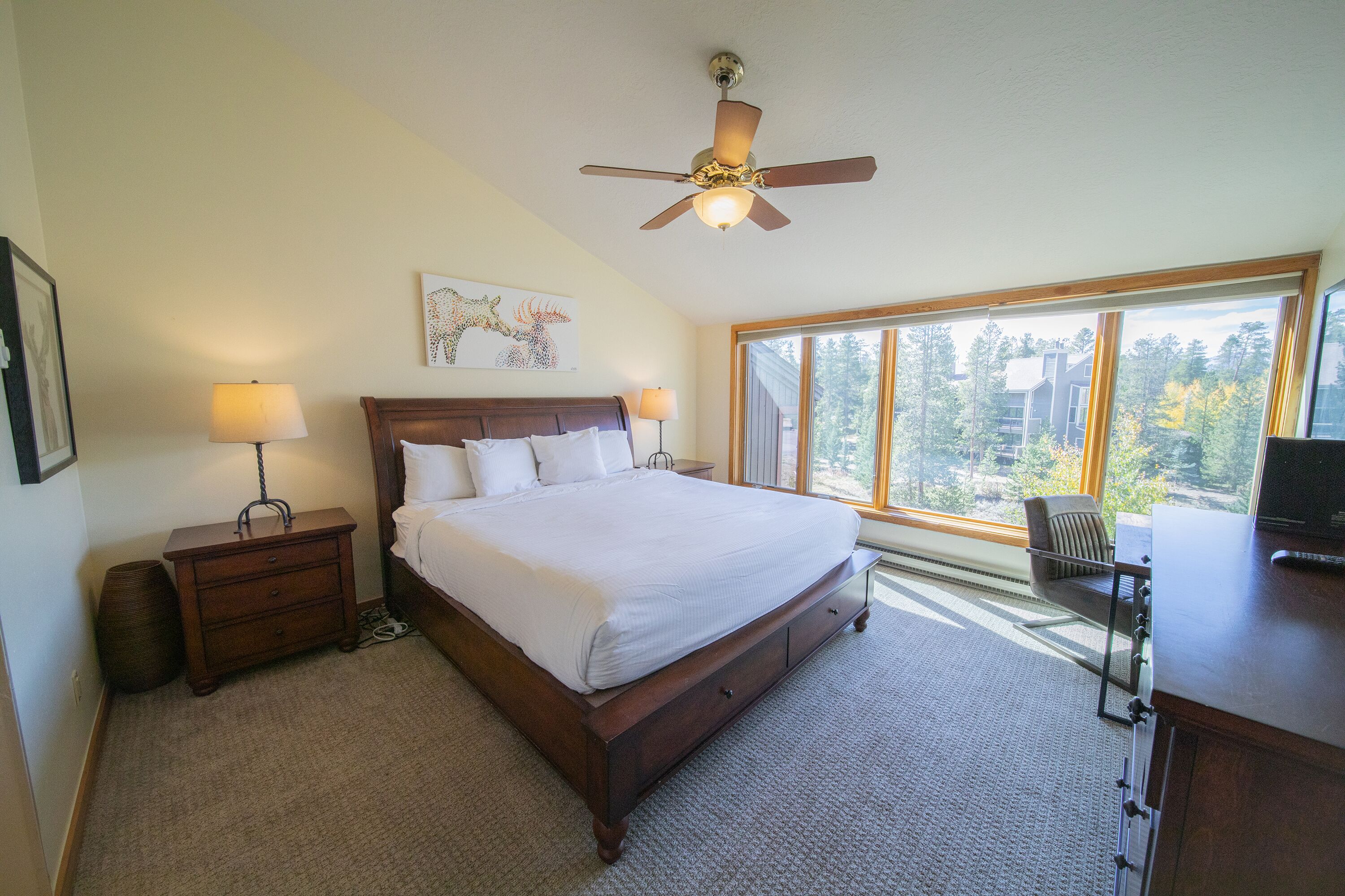 A bedroom with a large bed, two bedside tables with lamps, a ceiling fan, a desk with a chair, and a large window offering a view of trees and buildings.