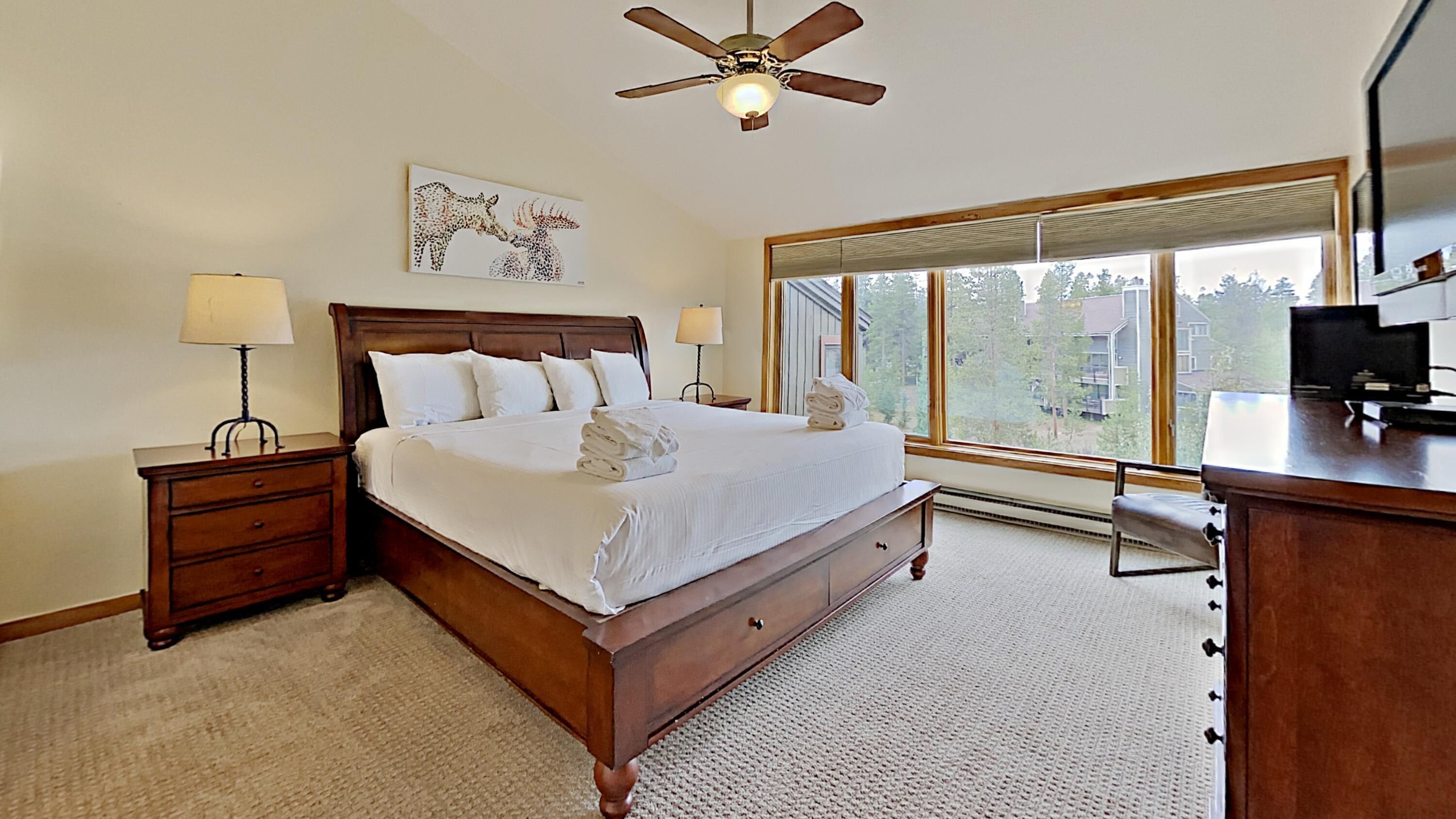 A bedroom with a large bed, two bedside tables, a ceiling fan, a window with a view, and a desk with a chair.