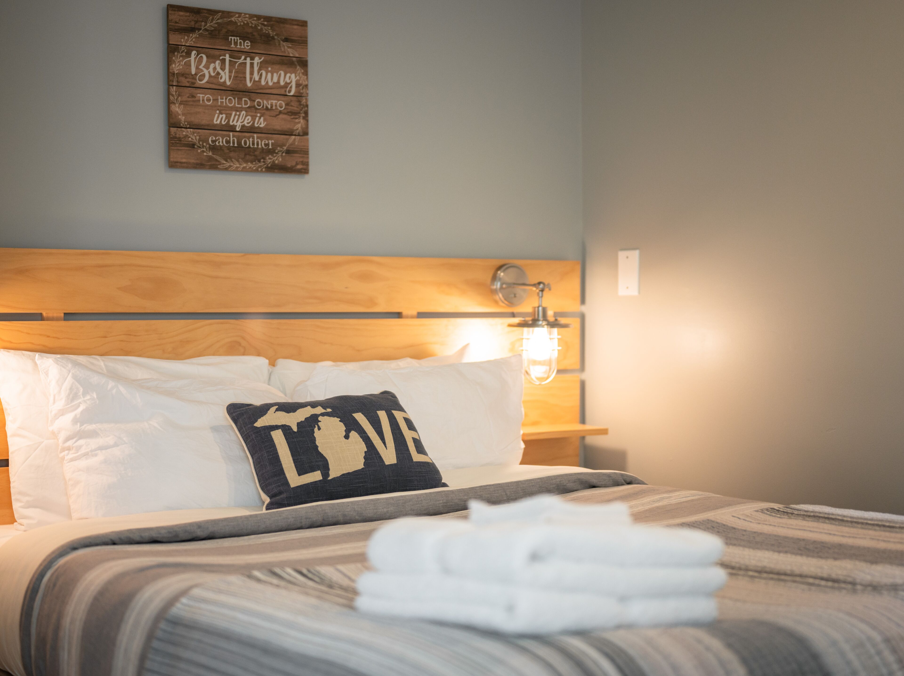 A neatly made bed with white linens and a pillow with "LOVE" on it. There is a wall-mounted sign with a quote.