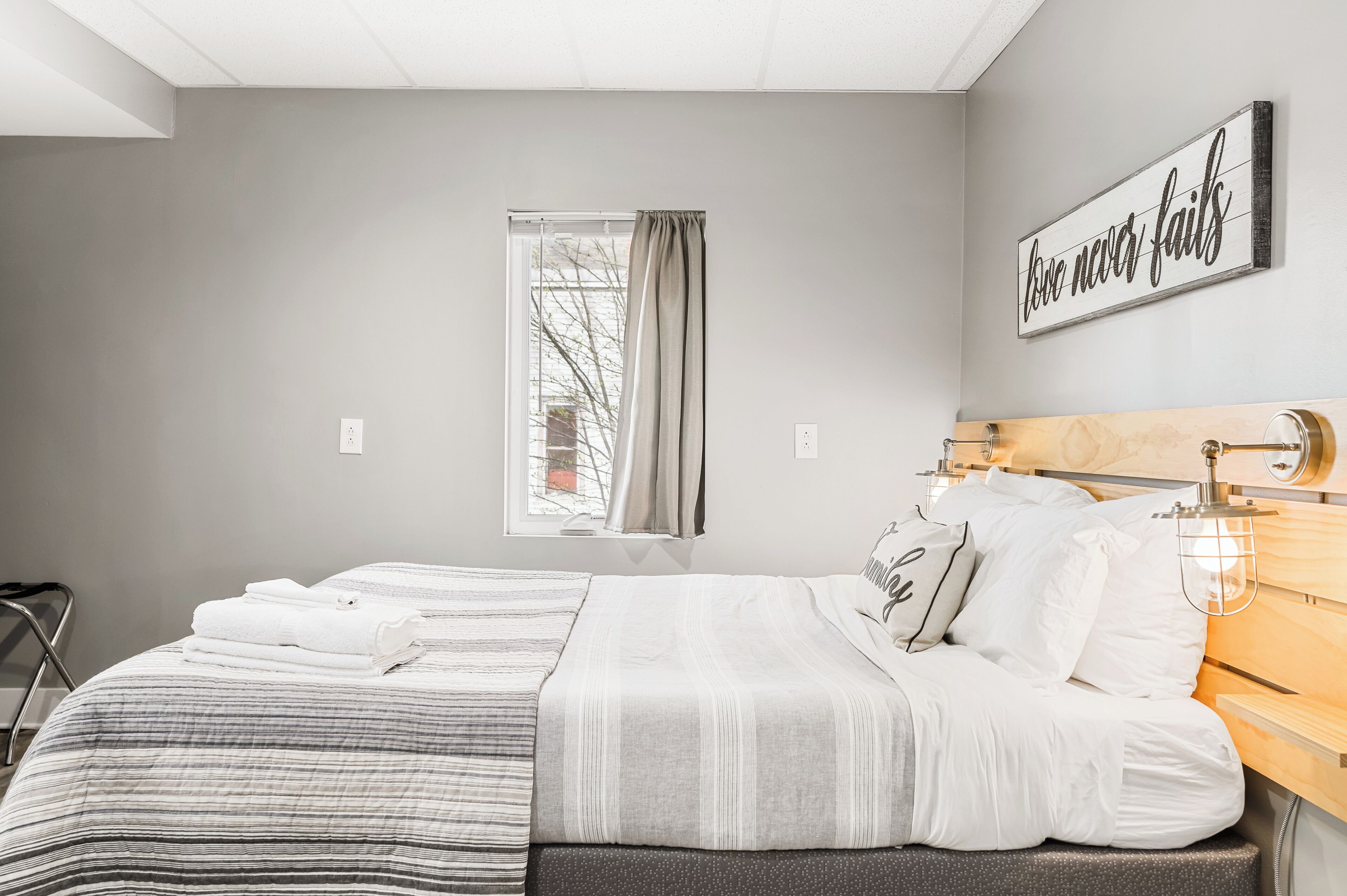 A neatly made bed with white linens, a window with curtains, and a motivational quote on the wall.