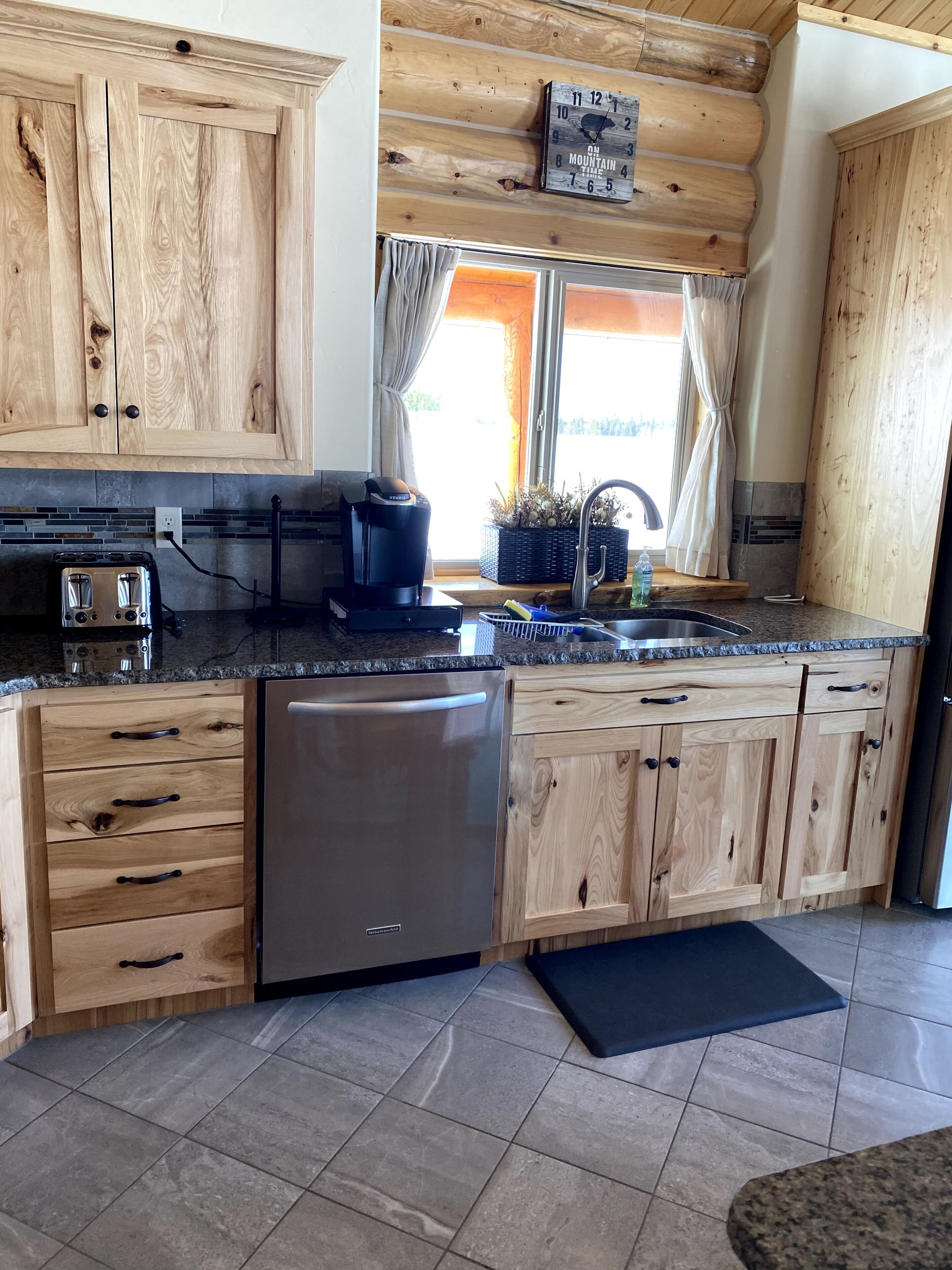A kitchen with wooden cabinets, a stainless steel dishwasher, a coffee maker, and a window with curtains.
