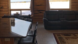 L'intérieur d'une cabane comprend une table à manger, des chaises, un canapé et de grandes fenêtres donnant sur un paysage enneigé.