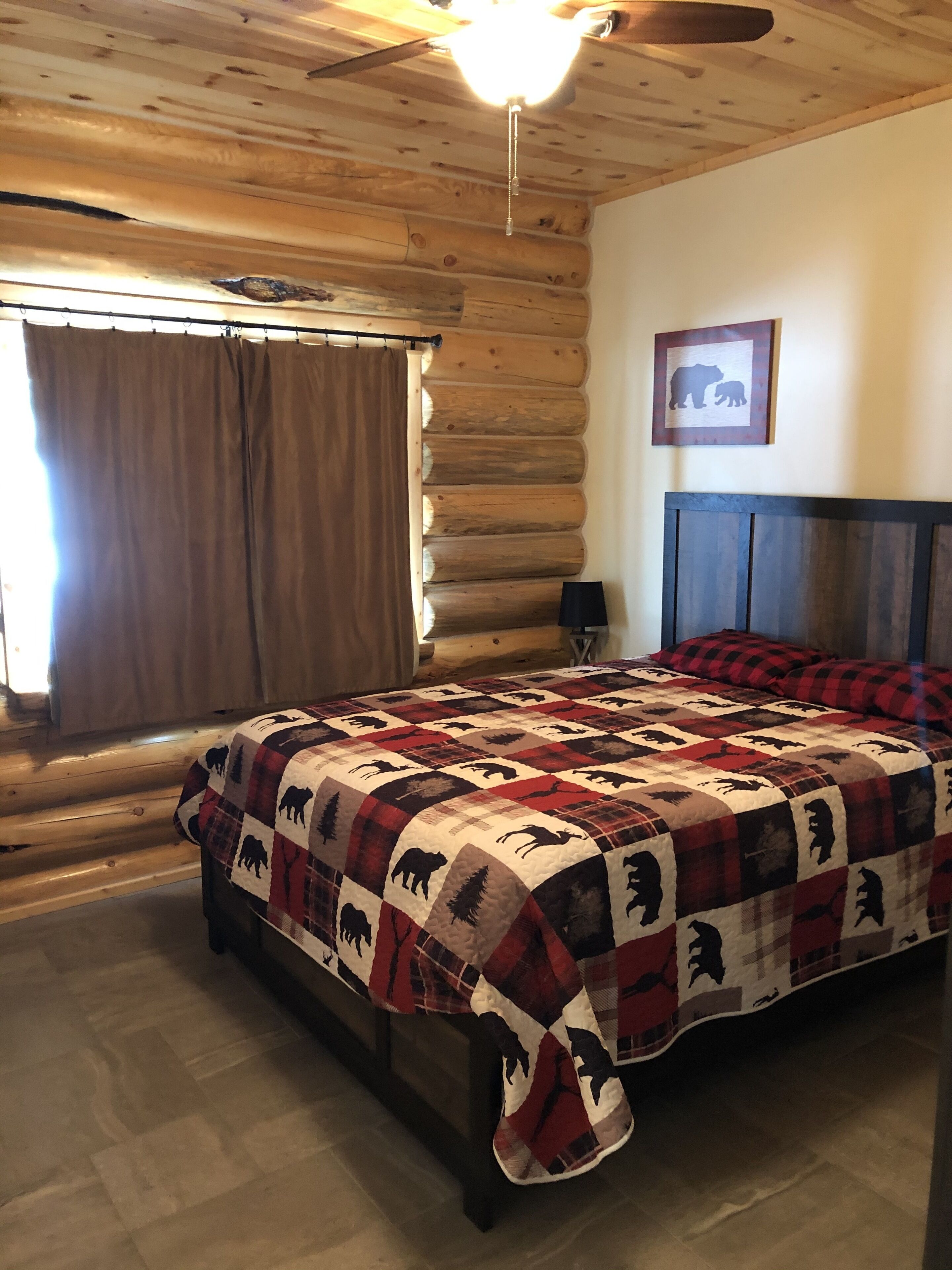 A log cabin bedroom with a bed, a ceiling fan, a window with curtains, and a bear silhouette picture on the wall.