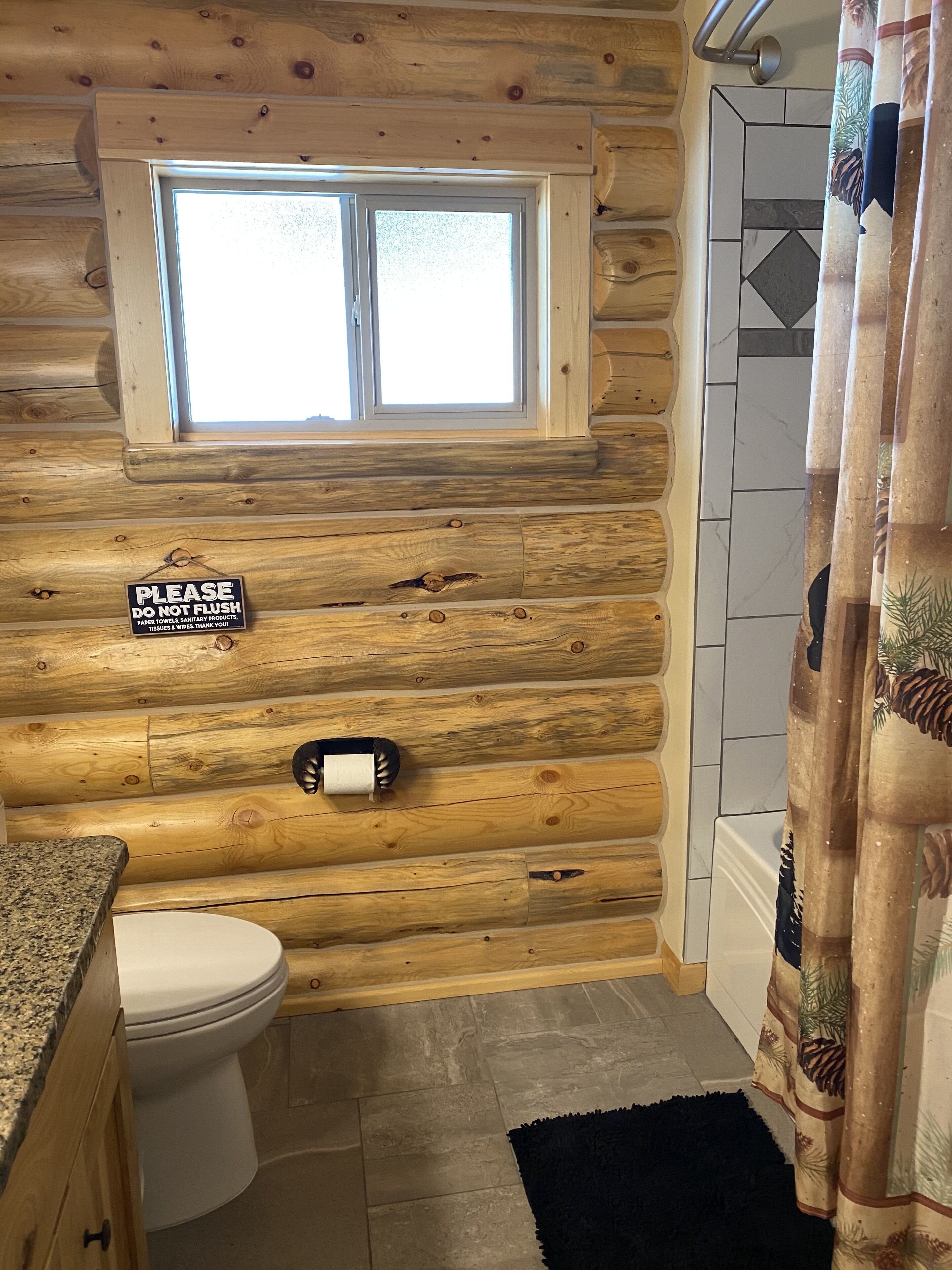 A bathroom with a wooden log wall, a toilet, a sink, a window, and a shower curtain.