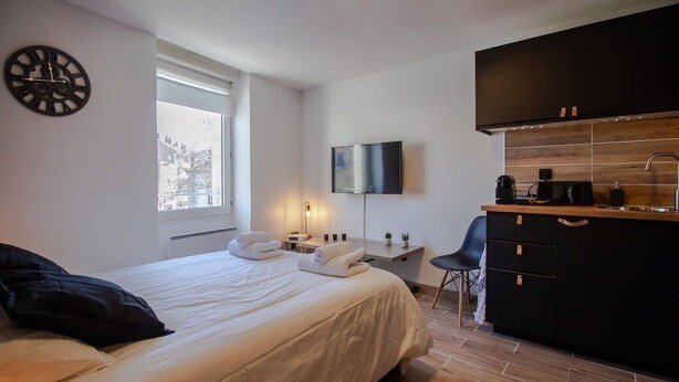 A modern hotel room with a bed, a desk with a flat-screen TV, a kitchenette, and a large window. - THE SQUARE ROOT (Dijon)