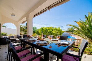 Un área de comedor al aire libre con vista a la piscina, mobiliario de mimbre y cielo despejado.