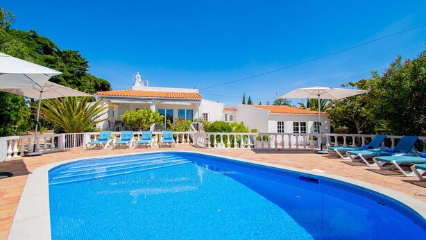 A swimming pool with lounge chairs and umbrellas, surrounded by a white fence and a building with a tiled roof. - Montenegro Superbly Located Family Vila With Game Room & Pool (Carvoeiro)