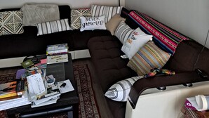Uma sala de estar com sofá seccional, mesa de centro com livros e revistas, e tapete estampado.