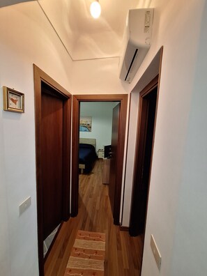 A narrow hallway with wooden flooring, a carpeted area, and a bed in the background.