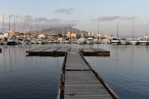 A marina with numerous boats docked, a wooden pier leading into the water, and a cityscape with buildings and mountains in the background.