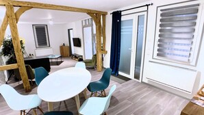 Ein modernes Wohnzimmer mit einem runden Tisch, vier Stühlen, Holzbalken, einer Schiebetür aus Glas und einem großen Fenster mit Jalousien.