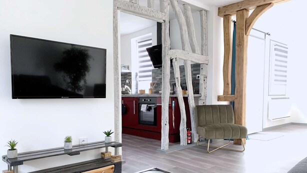 Ein modernes Wohnzimmer mit einem an der Wand befestigten Flachbildfernseher, einem Couchtisch aus Holz mit geometrischem Design, einer roten Küchenzeile mit Herd und Backofen sowie einem grünen Sessel.