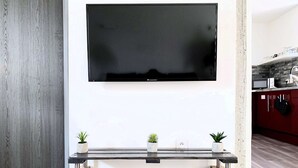 Ein modernes Wohnzimmer mit einem an der Wand befestigten Flachbildfernseher, einem Glastisch mit Holzbeinen und drei kleinen Topfpflanzen.