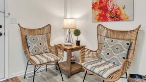 A corner room with two wicker chairs, a small table with a lamp, and a painting on the wall. - Walk Downtown | Parkside | City View | Beautifully Remodeled (Ann Arbor)