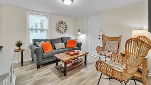A living room with a grey sofa, orange cushions, a wooden coffee table, and wicker chairs. - Walk Downtown | Parkside | City View | Beautifully Remodeled (Ann Arbor)