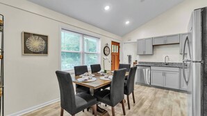 A modern kitchen with grey cabinets, a stainless steel refrigerator, and a dining area with a wooden table and grey chairs. - Walk Downtown | Parkside | City View | Beautifully Remodeled (Ann Arbor)