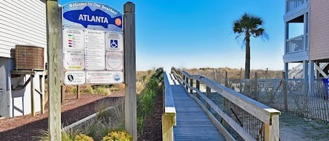 Une promenade en bois mène à une zone de plage où se trouve un panneau affichant les règles de la plage et des informations de sécurité.