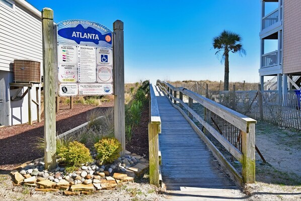 A wooden boardwalk leading to a beach area with a signboard displaying beach rules and safety information.
