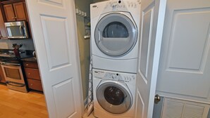 A laundry room with two stacked front-loading washing machines, a microwave, and a stove in the adjacent kitchen area. - Blue Heaven - Walk to Beach & Boardwalk, Ocean and Sunset Views, Dog-Friendly (Carolina Beach)