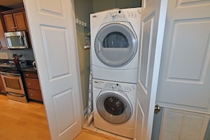 A laundry room with two stacked front-loading washing machines, a microwave, and a stove in the adjacent kitchen area. - Blue Heaven - Walk to Beach & Boardwalk, Ocean and Sunset Views, Dog-Friendly (Carolina Beach)