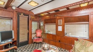 A train car bedroom with a television, a red armchair, a table with a bowl, and wooden paneling.