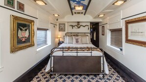 A bedroom with a bed centered in the room, flanked by windows with blinds, a ceiling with a cloud design, and framed artwork on the walls.