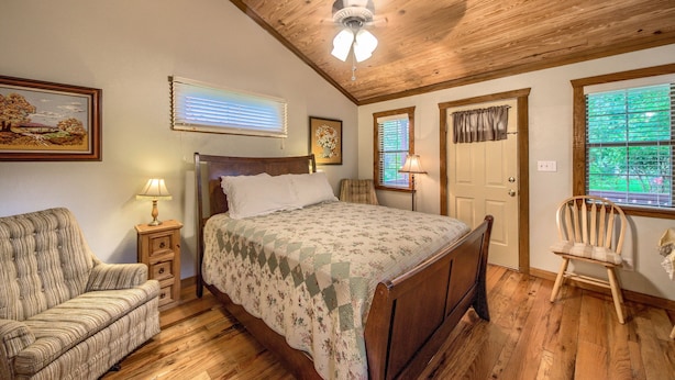 A bedroom with a wooden ceiling, a bed with a floral bedspread, a chair, a nightstand with a lamp, and a window with blinds. - 1 mile to downtown - Short Drive - Jacuzzi Bath (Fredericksburg)