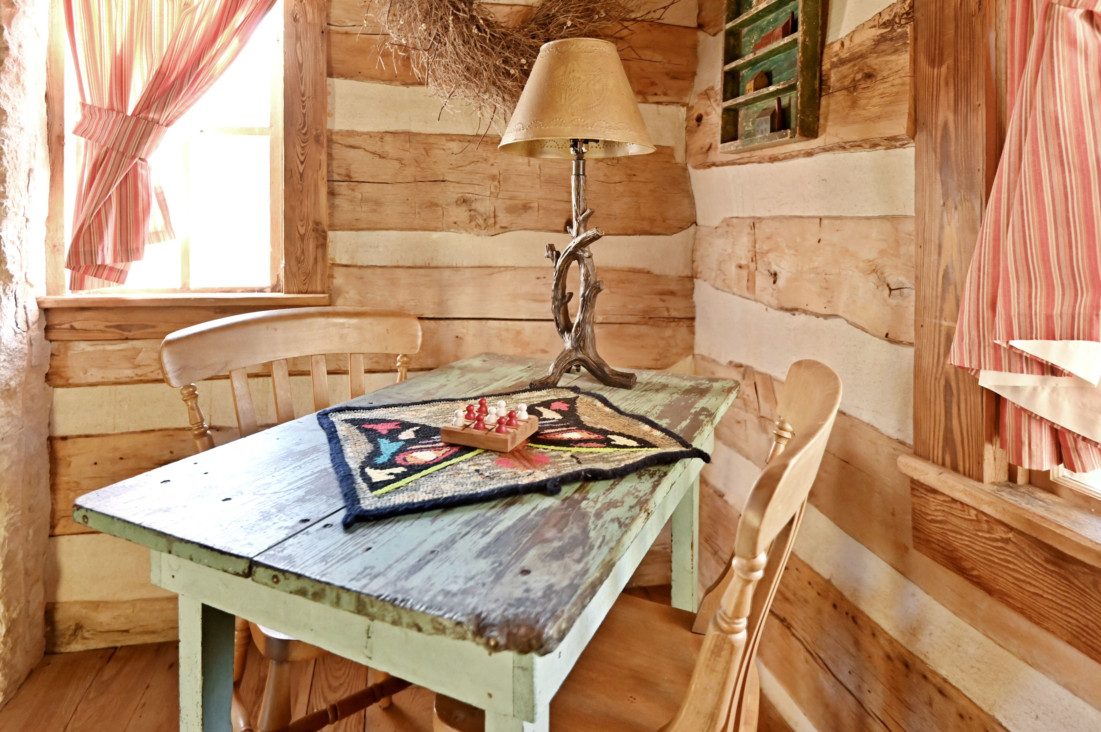 A rustic wooden cabin interior with a table, chairs, a lamp, and a window with curtains.