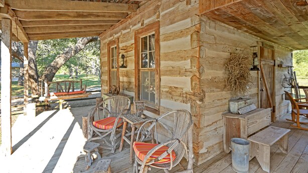 Une cabane rustique en rondins de bois, dotée d’un porche avec des fauteuils en osier ornés de coussins rouges, une table en bois et un luminaire suspendu.
