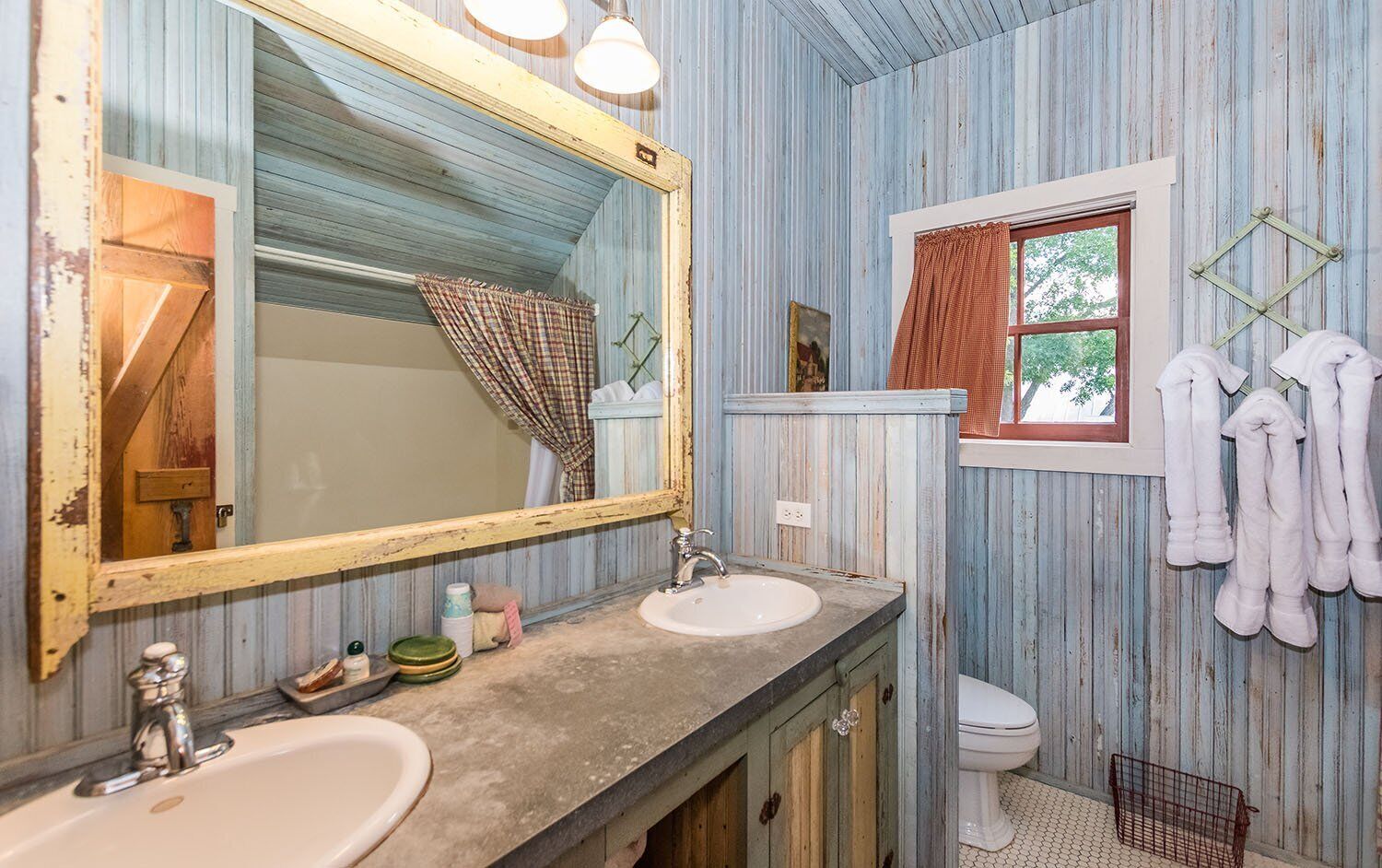 A bathroom with wooden walls, a large mirror, two sinks, a toilet, and towels hanging on hooks.