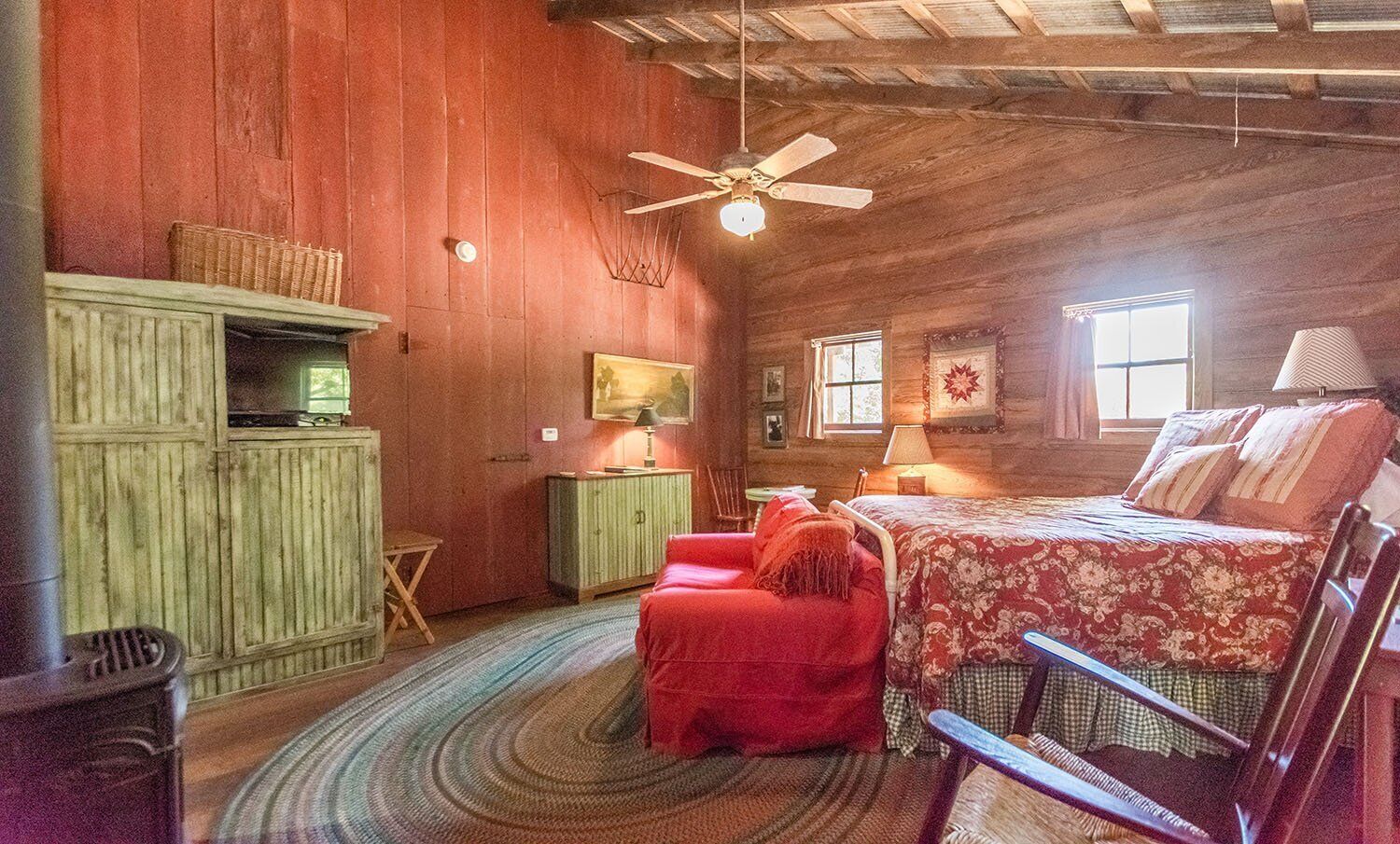 A rustic room with a bed, a red chair, a small table, a cabinet, and a ceiling fan.