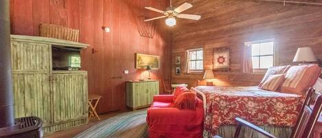 A rustic room with a bed, a red chair, a small table, a cabinet, and a ceiling fan.