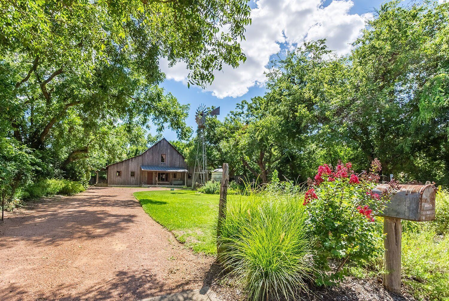 A rustic barn with a dirt path leading to it, surrounded by lush greenery and flowering plants.