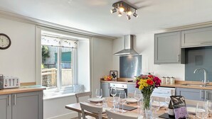Une cuisine moderne avec des meubles gris, un coin repas avec des chaises blanches, une grande fenêtre et un luminaire au plafond.