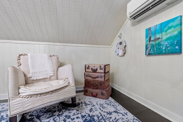 A room with a white textured ceiling, a white wall with a painting, a white chair with a blanket, and stacked vintage suitcases. - Charming Private 2-Story Cottage - Romantic Getaway - Close to Downtown (Fredericksburg)