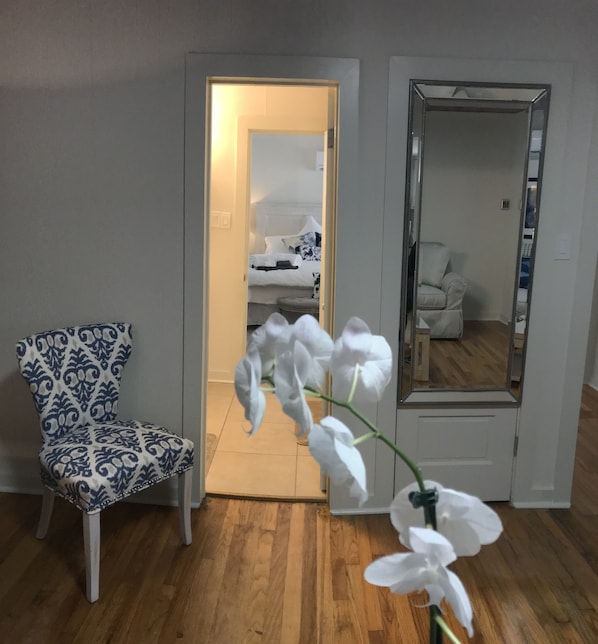 Una habitación con una silla estampada, una orquídea blanca, un espejo y una puerta que da a otra habitación con una cama y una silla.