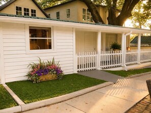 Una casa de un piso con fachada blanca, un porche cubierto y una valla de piquetes blancos, junto a un pequeño jardín con flores.
