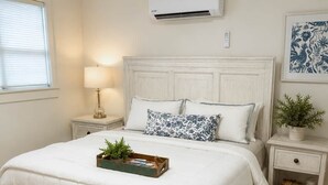 A bedroom with a large bed, a bedside table with a lamp, a small potted plant, and a wall-mounted air conditioner.
